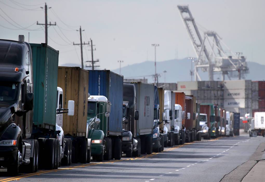 Row of diesel semi-trucks queued near Port of Oakland cranes — present-day industrial pollution burden in West Oakland